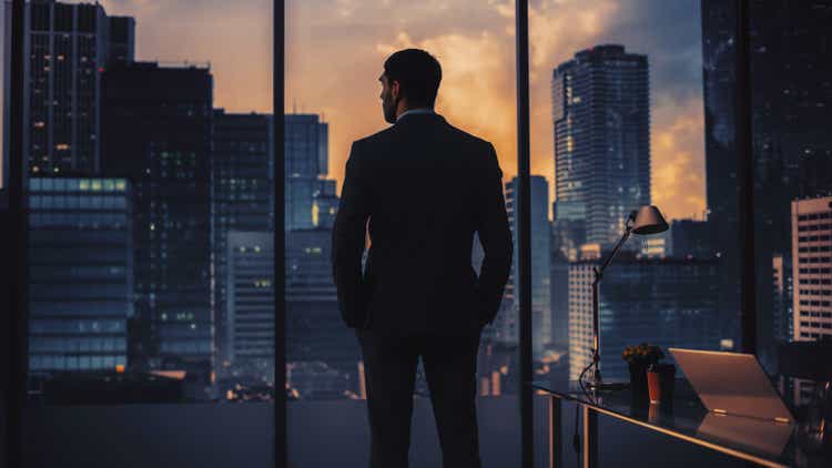 Thoughtful Young Businessman in a Perfect Tailored Suit Standing in His Modern Office Looking out of the Window on Big City in the Evening. Successful Finance Manager Planning Project Strategy.