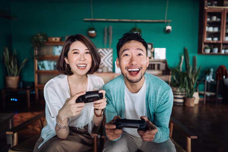 Happy young Asian couple sitting on the sofa in the living room, having fun playing video games together at home