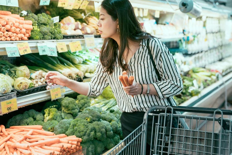 sivukuva aasialainen urbaani nainen, jolla on porkkanoita kärryssä, nojaa lähelle katsomaan hintalappua ostaessaan tuotteita paikallisesta ruokakaupasta.