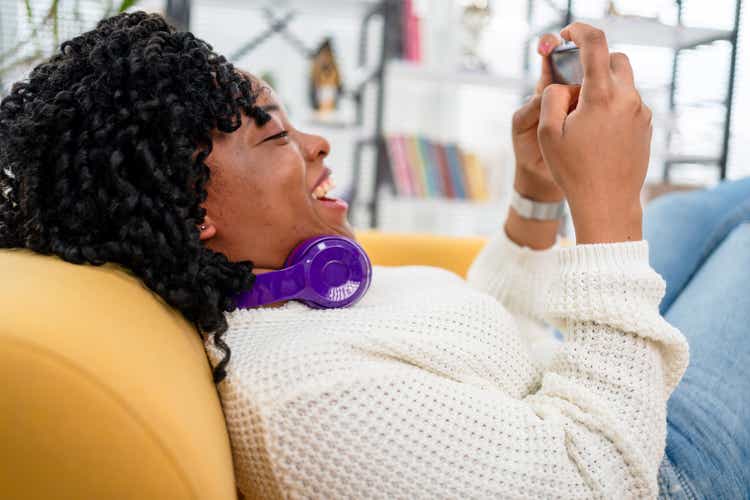 Happy black girl gamer wear headphone competition play video game online with smartphone in living room at home.