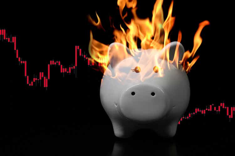 White piggy bank on fire on a very dark surface with descending red candle chart behind. Concept for economic crisis, inflation, devaluation, financial loss, devalued savings, market problems.