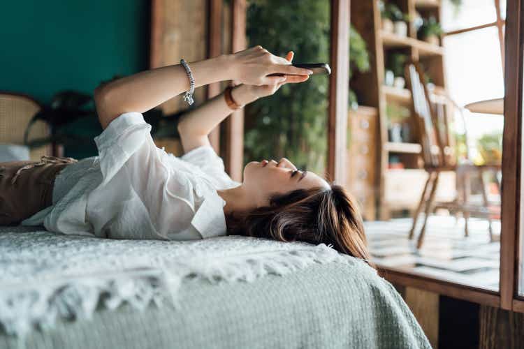 Relaxed young Asian woman shopping online with smartphone while lying in bed. Youth culture. Technology in everyday life