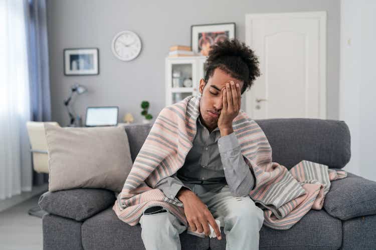 Young african american man, sitting on the sofa at home, having a fever and headache and touching his forehead to check his temperature