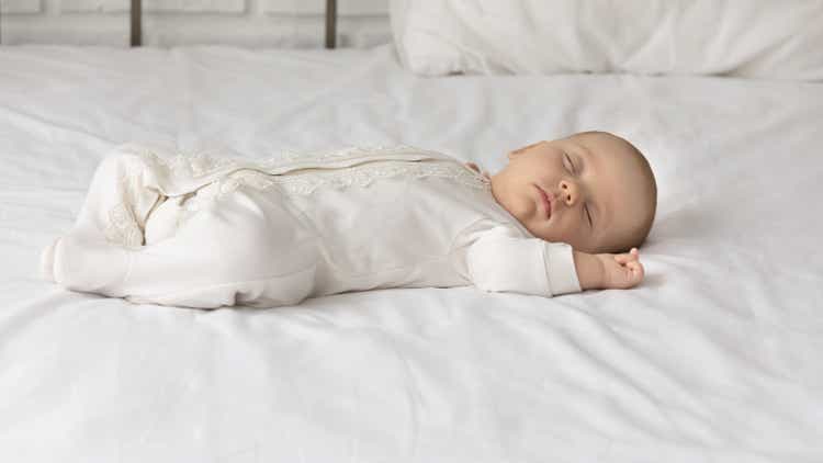 Cute baby in white bodysuit sleeping on comfortable bed