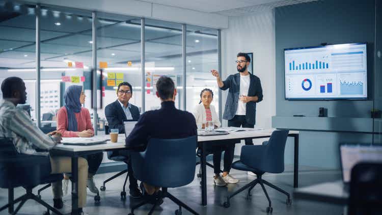 Diverse Modern Office: Businessman Leads Business Meeting with Managers, Talks, uses Presentation TV with Statistics, Infographics. Digital Entrepreneurs Work on e-Commerce Project.