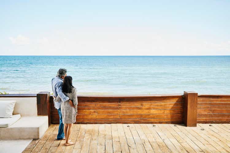 Wide shot of senior couple embracing and enjoying view on deck of luxury suite at tropical resort