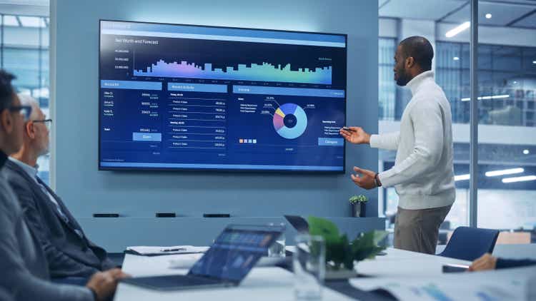 Diverse Modern Office: Motivated Black Businessman Leads Business Meeting with Managers, Talks, uses Presentation TV with Statistics, Charts, Big Data. Digital Entrepreneurs Work on e-Commerce Project