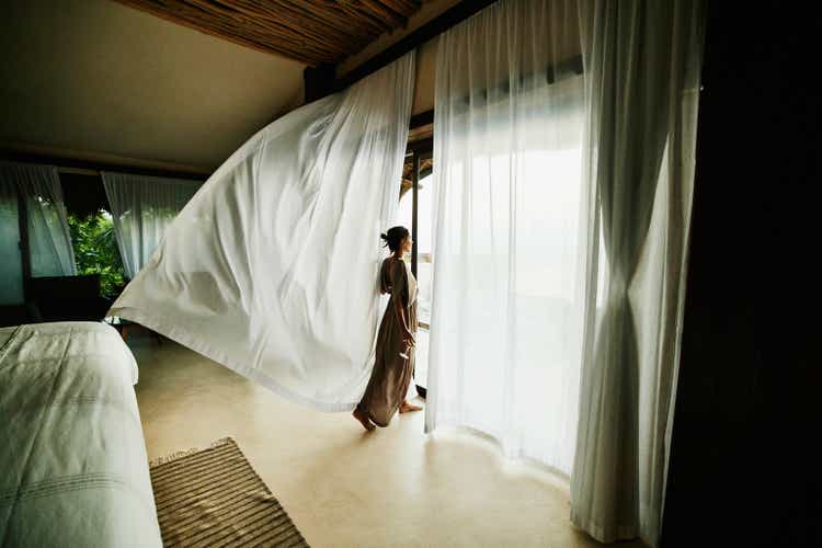 Wide shot of woman standing in luxury hotel suite looking at view with curtains blowing in wind