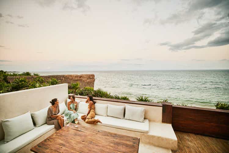 Wide shot of smiling female friends relaxing on deck of luxury hotel suite at sunset