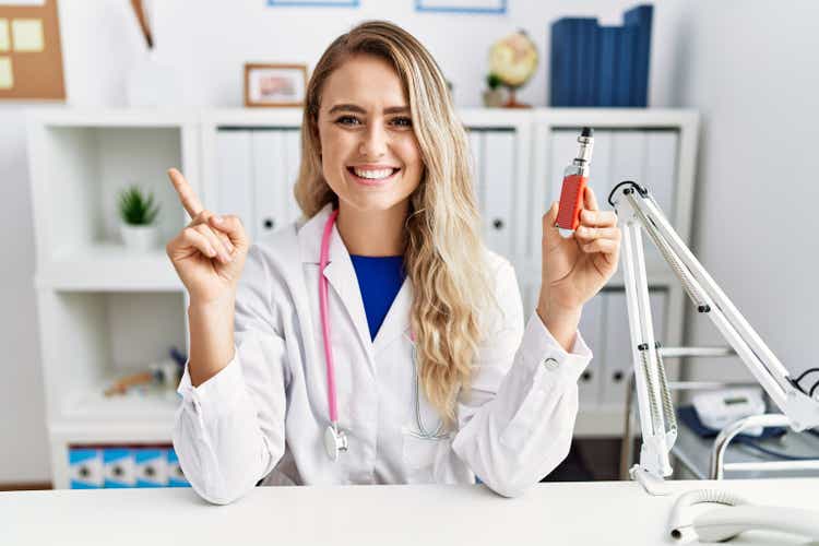 Young beautiful doctor woman holding electronic cigarette at the clinic smiling happy pointing with hand and finger to the side
