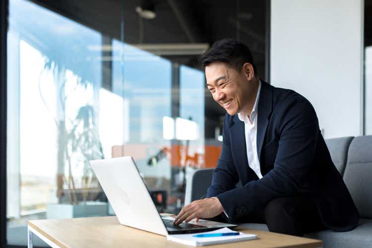Senior Asian business man working on laptop online, smiling and rejoicing, happy boss business owner working in office