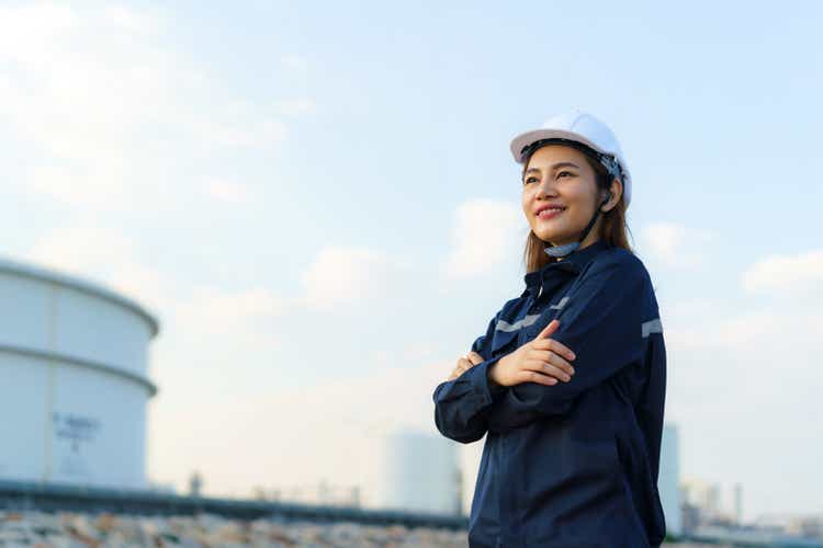 Asian woman engineer arm crossed and smile with confident looking forward to future with oil refinery plant factory in background.