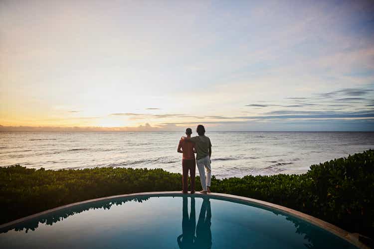 Wide shot of embracing gay couple watching sunrise while standing at edge of pool at luxury tropical villa overlooking ocean