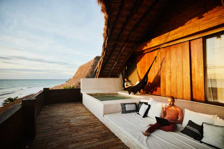 Wide shot of man drinking coffee and working on laptop while partner relaxes in hammock on deck of luxury tropical beach villa at sunrise