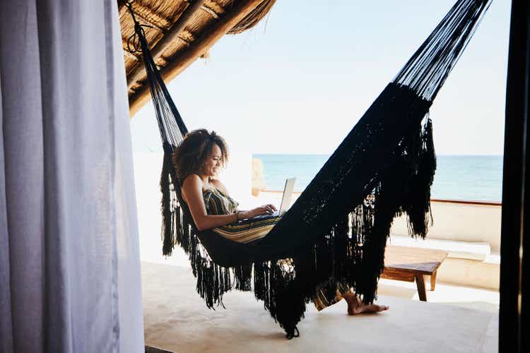 Wide shot of woman working on laptop while relaxing in hammock on deck of luxury tropical villa overlooking ocean