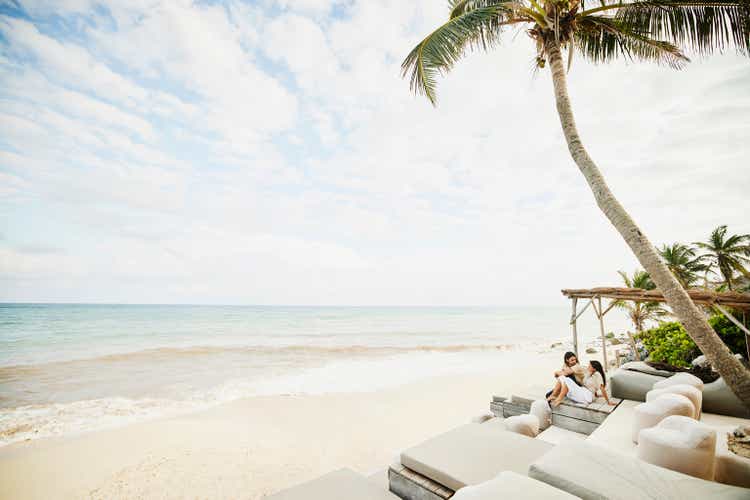 Extreme wide shot of smiling couple relaxing in tropical resort lounge area overlooking beach