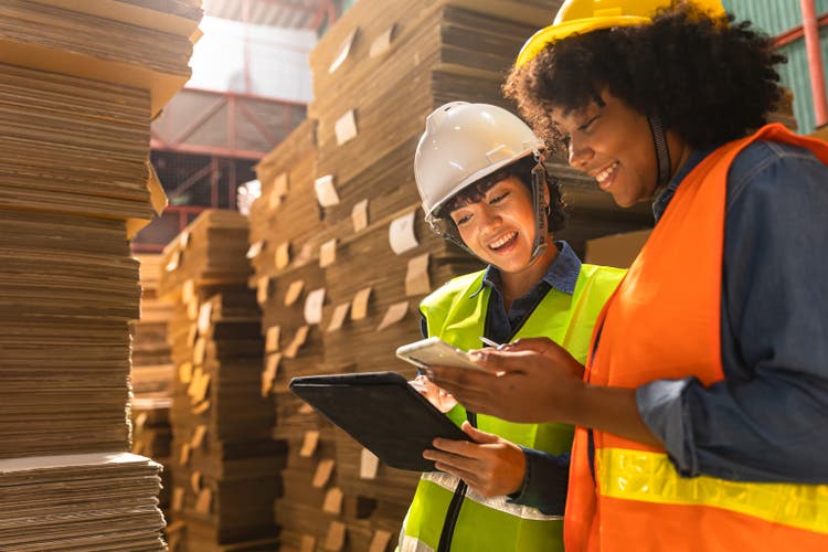 Engineer asian and african woman wearing safety helmet and vest holding clipboard and take note on the paper in the automotive part warehouse.Products and corrugated cardboard. Factory for the manufacture and processing of paper.