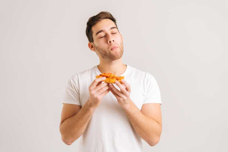 Portrait of satisfied young man with enjoying eating delicious slice of pizza, with closed eyes from pleasure on white isolated background.