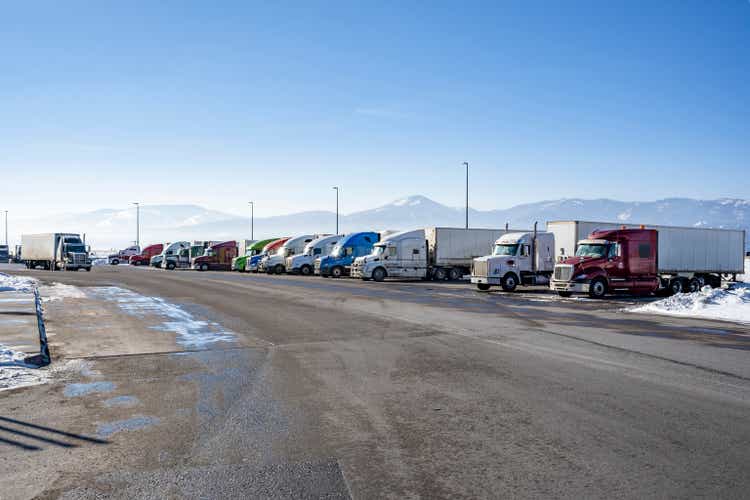 Big rig semi trucks with semi trailers standing in row on the snow and icy truck stop parking lot at winter season