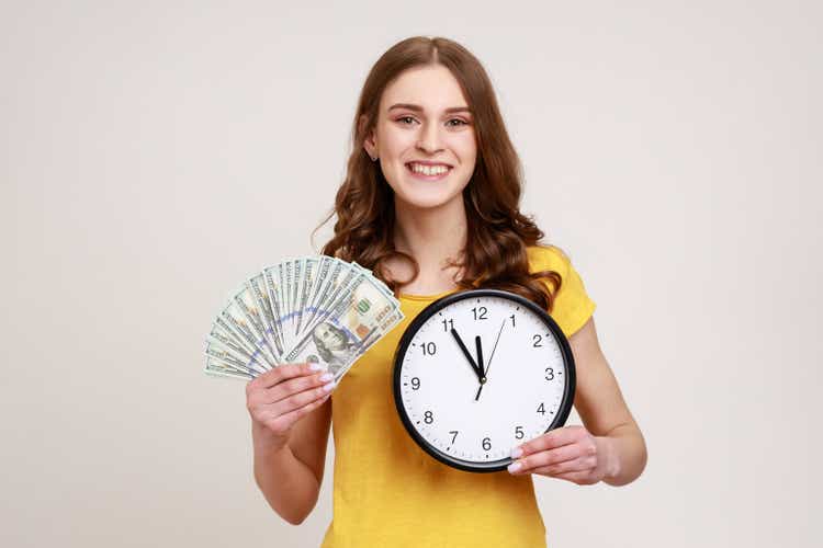 Time to make money. Happy smiling young woman showing dollar cash and wall clock on camera, hourly payment, expressing positive emotions.