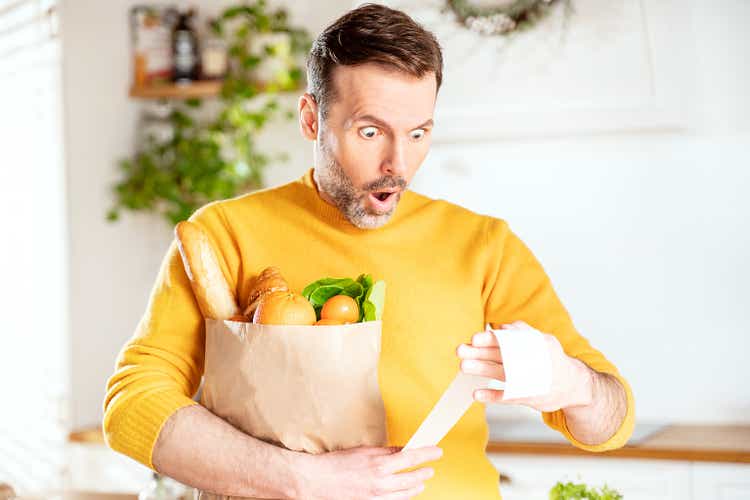 Surprised man looking at store receipt after shopping, holding a paper bag with healthy food. Guy in the kitchen.