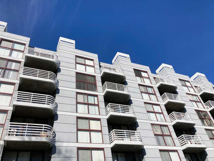 Exterior view of typical multifamily mid-rise residential apartment building with balconies and identical windows blinds under blue sky