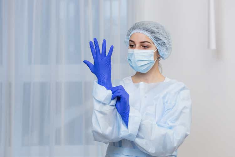 Young female doctor prepares for surgery, wears blue surgical gloves, in a white coat and mask, looks at the camera