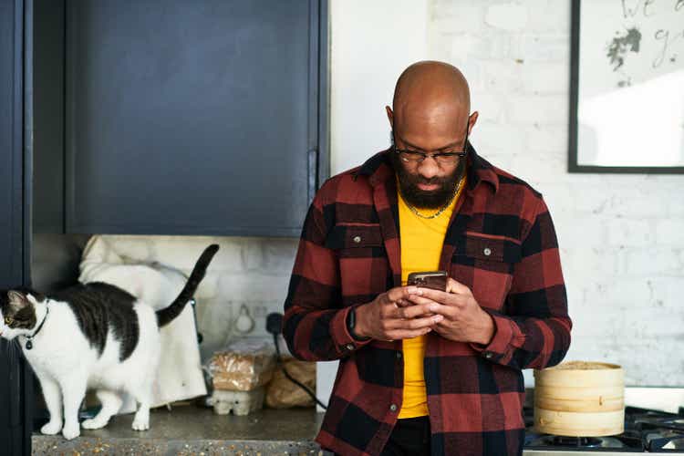 Mid adult black entrepreneur checking mobile phone with cat on kitchen worktop