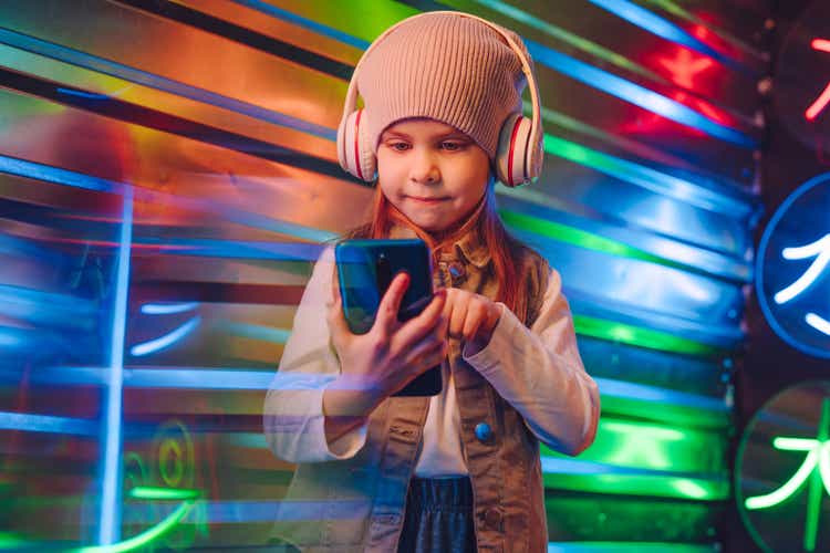 Little girl wearing headphones using smartphone on the colorful neon