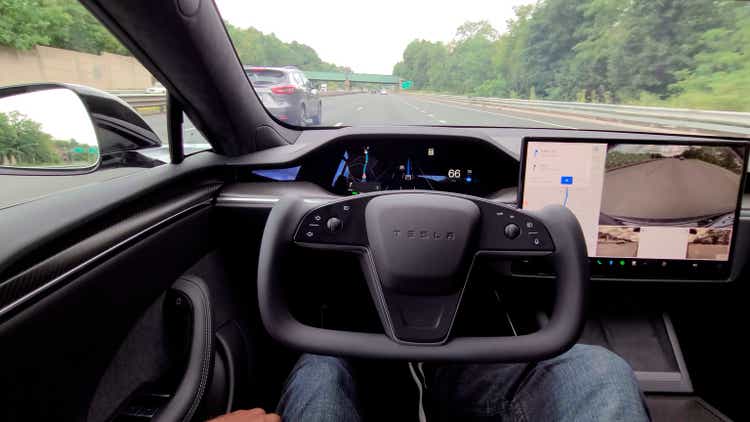 Close-up of a patterned Tesla Model S, driving down a highway on autopilot.  A man sits with his hands on his feet.