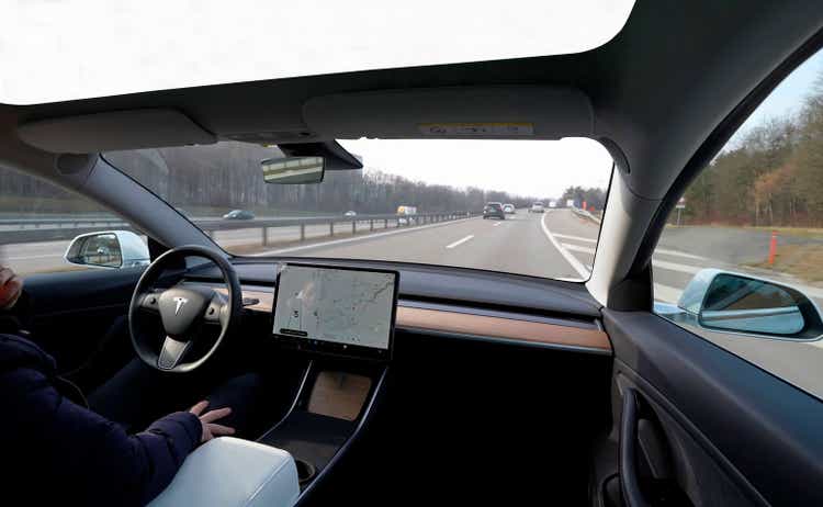 Close-up of a Tesla Model 3, with a glass roof. driving down a highway on autopilot. And a man inside, resting, with one hand on his face and the other on his leg.