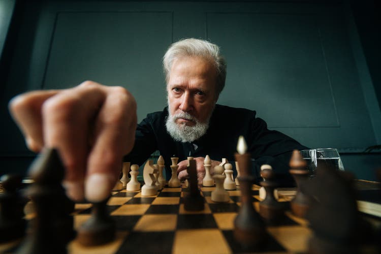 Close-up low-angle view of serious gray-haired mature male performing move with pawn piece on wooden chessboard, looking at camera.