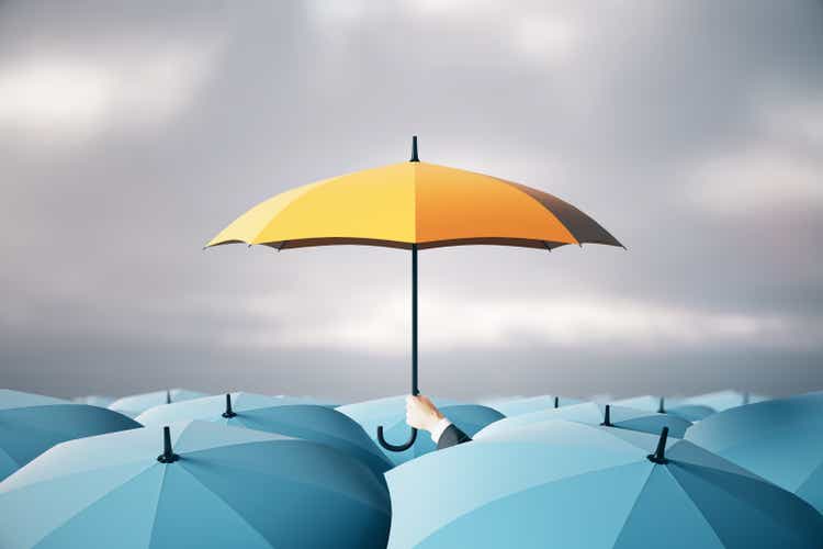 Businessman hand holding yellow umbrella over crowd on blurry dull sky background. Risk, protection and safety concept.