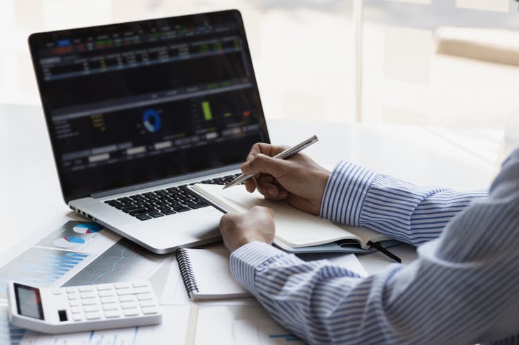 A businessman writes on a notepad while working on a laptop and is using accounting software for account analysis and auditing.