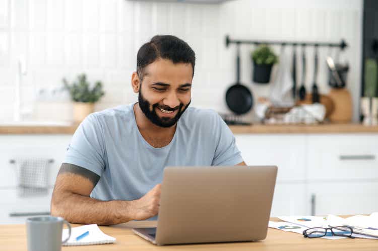 Positive handsome successful indian or arabian man, freelancer, designer, IT specialist, sitting at the table in the kitchen with a laptop, working online on a new project, smiling happily
