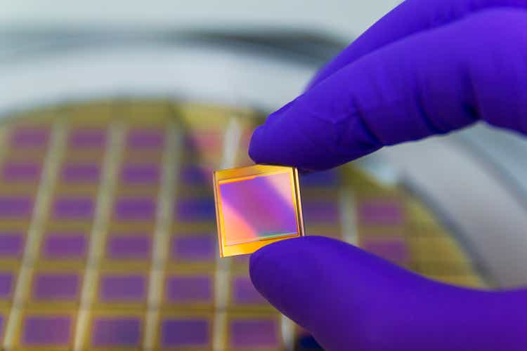 Hand in blue color glove holding microchip photo sensor matrix. On the background is diced silicon wafer with microchips. Focus on chip.