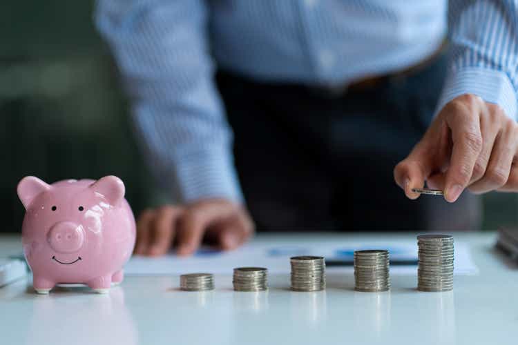 Close-up of young Asian businessman putting coins on pile of coins with colored piggy banks beside financial planning ideas to use enough income to save money.