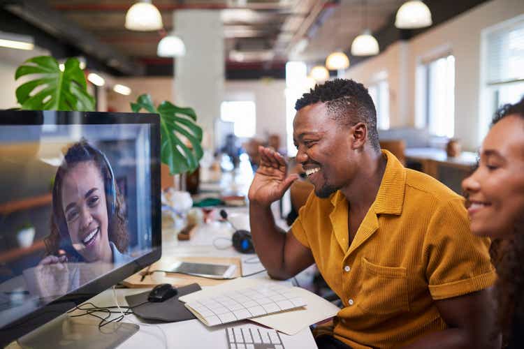 A Black man waves to his colleague on a video call from his office