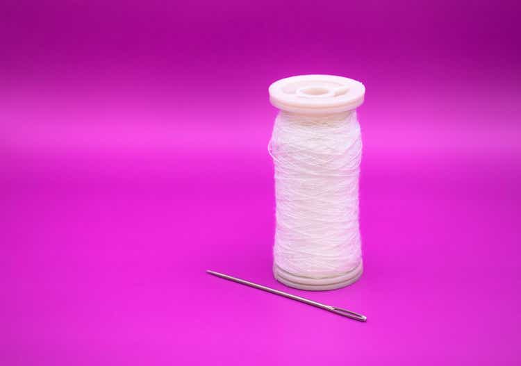 Spool of white colored thread accompanied by a sewing needle isolated on the purple background