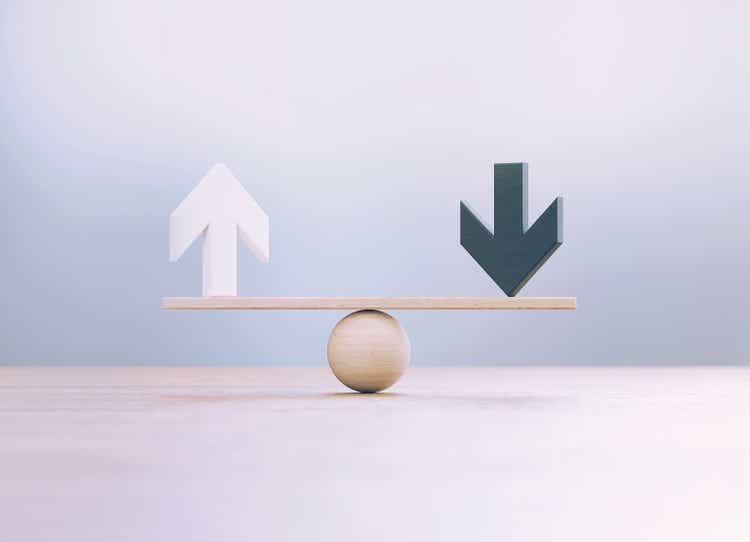 White And Black Wooden Arrow Symbols Sitting Over Wooden Seesaw Scale Before Defocused Background