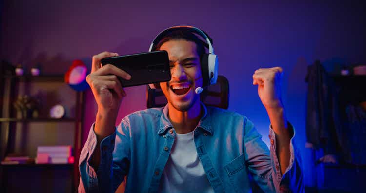 Happy asia man gamer wear headphone competition play video game online with smartphone colorful neon light in living room at night modern house.