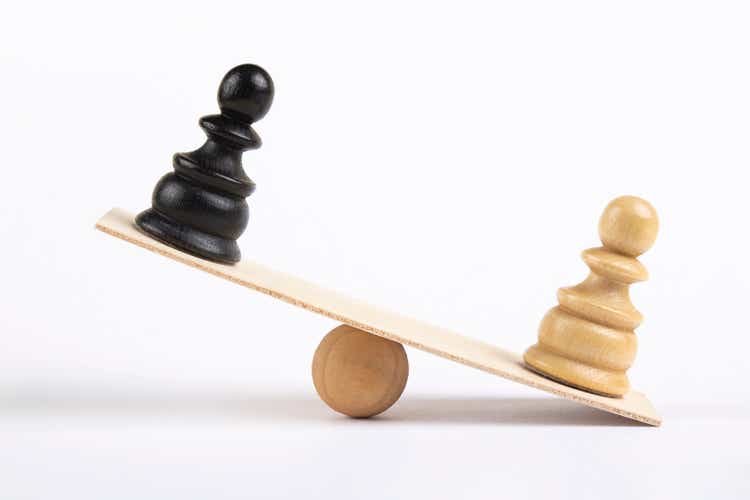 White and a black pawn chess piece stand on wooden seesaw