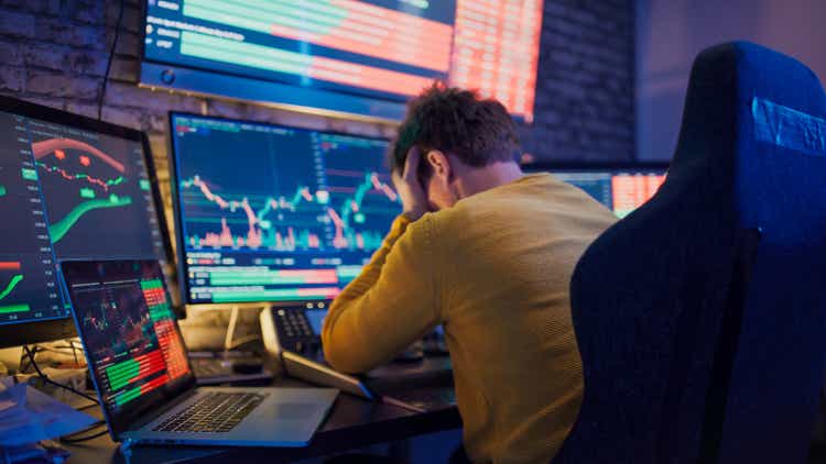 Worried man with head in hands sitting in front of stock market graphs on computer screens at desk