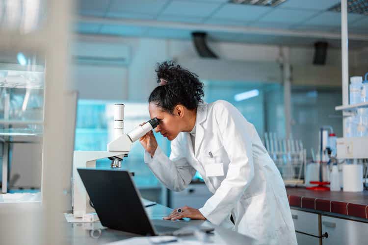 Female Scientist Looking Under Microscope And Using Laptop In A Laboratory