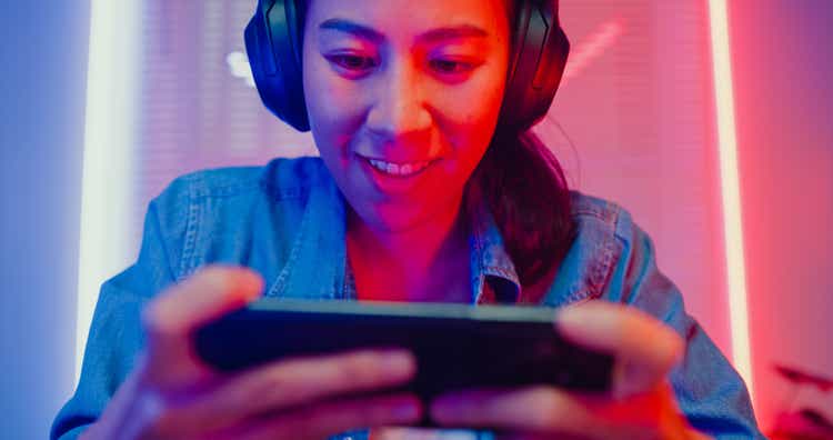 Close up of young Asian woman playing online smartphone video game and broadcast streaming live in neon lights living room at home.