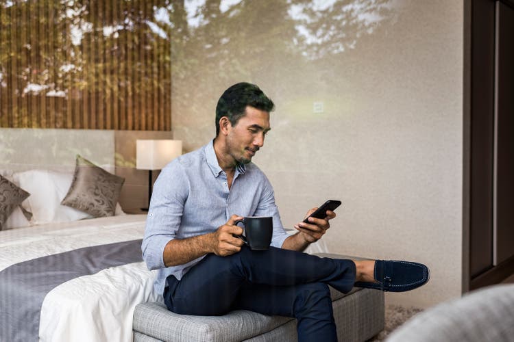 Young Asian businessman working from home, using smartphone, sitting on bed at bedroom window of modern house