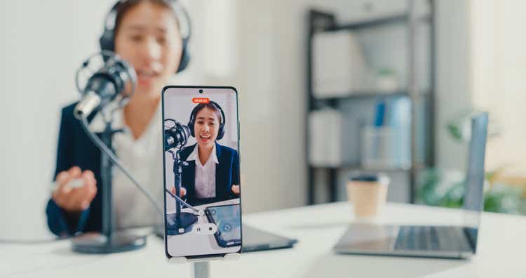 Close up of young Asian businesswoman recording content with smartphone and broadcasting a podcast on her laptop from studio office.