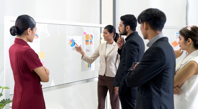 Young asian businesswoman in brown suit pointing finger at pie chart for business target, presented to group of investors. There was a note paper and document on the glass panel.