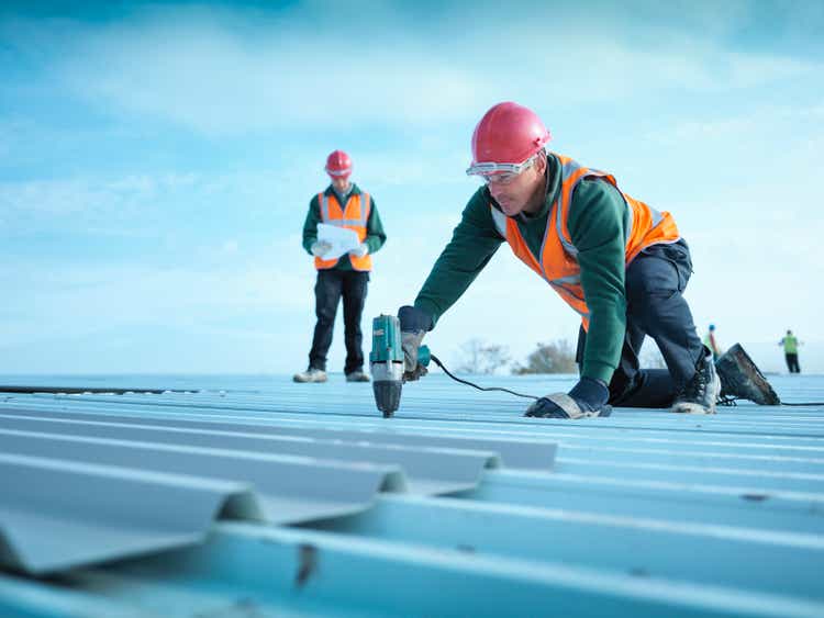 A construction foreman reads plans as a worker insulates a factory roof