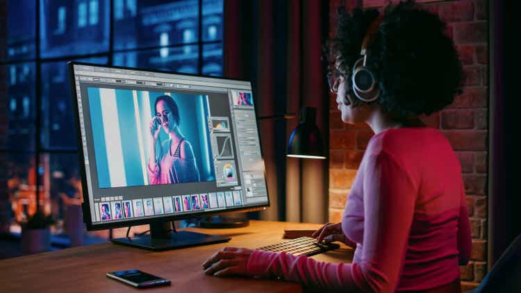 Young Creative Female in Headphones Using Computer in Stylish Loft Apartment in the Evening. Graphic Designer Working from Home, Editing Fashion Photo for Online Store. Urban View from Big Window.